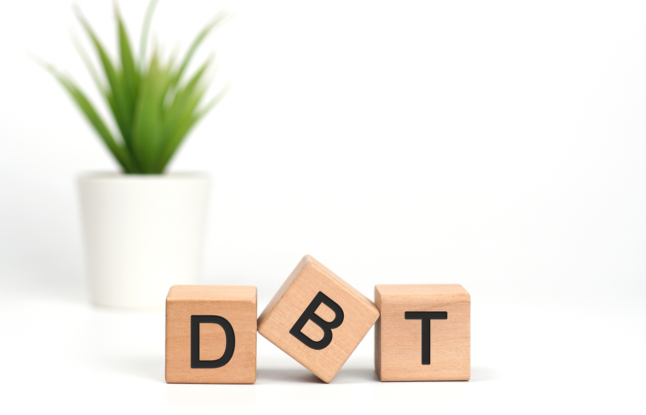 Wooden cubes with letters on white background. The words written is DBT Dialectical Behavior Therapy acronym with a pot of green plant.Psychological treatment concept, stress regulation.Copy space.