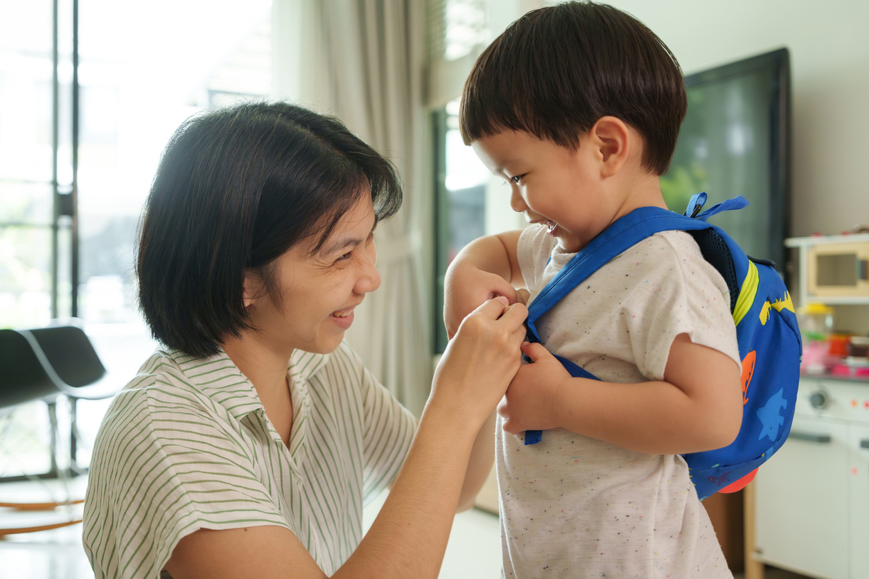 Smiling Asian mother adjusts blue backpack on happy young son. Close up in living room. Child laughs while mother ensures straps are secure. Parent child bonding, joy, and morning routine.